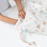 A child opens the Bumkins Wet Bag + Wet/Dry Bag Set: Palm Daze, reaching inside the palm tree-patterned bag on a white table.