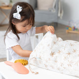 A young girl in a white bow opens a Bumkins Wet/Dry Bag: Palm Daze at a white table beside a toy shovel and sand toy.