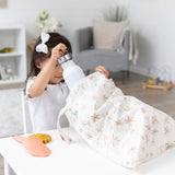 A child in a white shirt places a water bottle into a Bumkins Wet/Dry Bag: Palm Daze, with sand toys on the white table nearby.