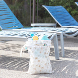 A poolside scene with blue lounge chairs and the Bumkins Palm Daze Wet/Dry Bag packed with sunscreen, water, and a towel.