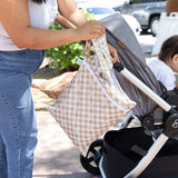 A person places clothes into a Bumkins Wet/Dry Bag: Sand Check attached to a stroller beside a sitting child outdoors on a sunny day.