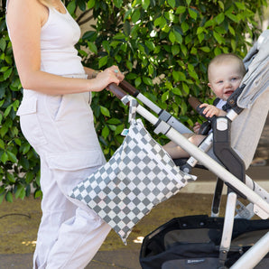 A person in a white outfit pushes a baby in a stroller with Bumkins Wet/Dry Bag: Charcoal Check attached. Green foliage in the background.
