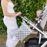 A person in a white outfit pushes a baby in a stroller with Bumkins Wet/Dry Bag: Charcoal Check attached. Green foliage in the background.