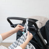 Hands attach the Bumkins Wet/Dry Bag: Charcoal Check to a stroller handle against a white brick wall.