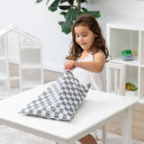 A young girl in a white dress sits at a white table, happily opening her Bumkins Wet/Dry Bag: Charcoal Check in a bright room.