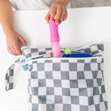 A child places a pink item into a Bumkins Wet/Dry Bag: Charcoal Check, featuring a zipper and waterproof fabric, on a white table.