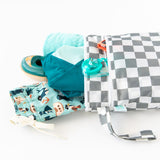 Bumkins Wet/Dry Bag: Charcoal Check with beach essentials on a white background: fun-pattern swim trunks, teal towel, swim shoes, and goggles.