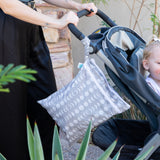A person pushes a stroller with a child. A Bumkins Wet/Dry Bag: Lunar Phase hangs from the handle, ideal for travel.