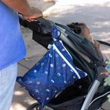 A person pushes a stroller with a child inside; a Bumkins Wet/Dry Bag: Evil Eye, made from waterproof fabric, hangs from the handle.