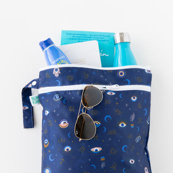 Bumkins Wet/Dry Bag: Evil Eye holds sunglasses, books, sunscreen, and a bottle on a white background.