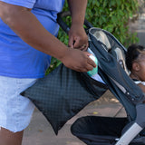 A person in blue places a bottle into a Roamio by Bumkins Wet/Dry Bag: Rich Black attached to a stroller with a child outdoors.
