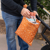 A person places clothes into a Bumkins Wet/Dry Bag: Grounded, attached to a stroller outdoors.