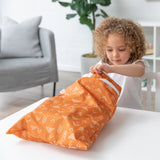A curly-haired child opens a Bumkins Wet/Dry Bag: Grounded at a white table. A sofa and plant add charm to the background.
