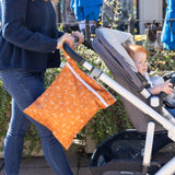 A woman pushes a stroller with a child outdoors; a Bumkins Wet/Dry Bag: Grounded hangs from the handle.