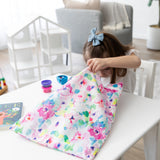 A young girl with a blue bow looks into her Bumkins Wet Bag + Wet/Dry Bag Set: Watercolor at a white table in a cozy room.
