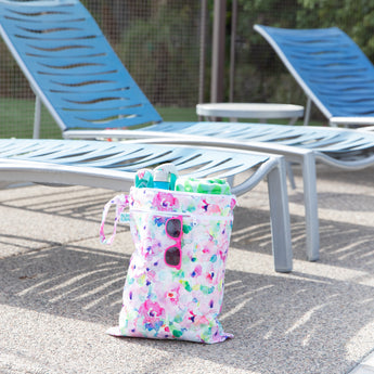 Bumkins Wet/Dry Bag: Watercolor holds pool items and pink sunglasses by blue lounge chairs on a sunny patio.