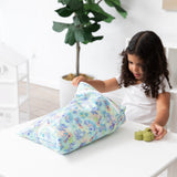 A young girl opens a Bumkins Wet/Dry Bag: Aloha Stitch while holding a green toy at a white table, with a potted plant in the background.