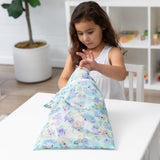 A girl in a white dress opens a colorful Bumkins Wet/Dry Bag: Aloha Stitch at a white table in a bright room with plants and shelves.