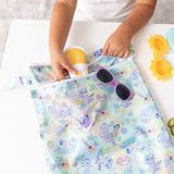 A child puts sunscreen and sunglasses in a colorful Bumkins Wet/Dry Bag: Aloha Stitch on a white table.