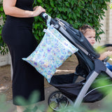 A person pushes a stroller with a Bumkins Wet/Dry Bag: Aloha Stitch hanging from the handle next to green foliage.