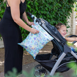 A smiling child sits in a stroller while a person unzips a Bumkins Wet/Dry Bag: Aloha Stitch attached to the handle.