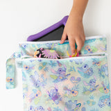 A child puts goggles, flower print clothes, and a purple tablet into a colorful Bumkins Wet/Dry Bag: Aloha Stitch on a white table.