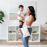 A woman smiles while holding a child and a Bumkins Wet/Dry Clutch: Palm Daze in a bright living room in front of shelves filled with toys.