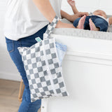 A person attends to a baby on a changing table while a Bumkins Wet/Dry Clutch: Charcoal Check hangs from their wrist.