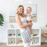 A smiling woman holds a baby and a Bumkins Wet/Dry Clutch: Charcoal Check in a bright room with toys on a shelf.