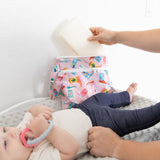 A person reaches for wipes from a Bumkins Wet/Dry Clutch: Boots on a changing table, sitting next to a baby clutching a teething ring.