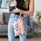 A woman holds a baby and a Bumkins Wet/Dry Clutch: Yoga Pals in a cozy living room with a sofa and plant.
