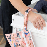 A person grips a changing table with a baby's leg visible, a Bumkins Wet/Dry Clutch: Yoga Pals hanging from their wrist.
