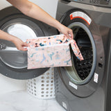 A person holds a Bumkins Wet/Dry Clutch: Yoga Pals in front of an open washing machine next to a white laundry basket.