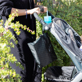 A person pushes a stroller with a cup and a Bumkins Wet/Dry Clutch: Rich Black amidst lush green foliage.