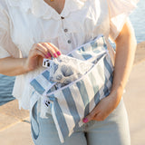 A woman in a white blouse removes a Roamio by Bumkins Reusable Snack Bag, Small 2 Pack: Coastal from a striped bag near water.