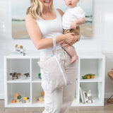 A mom smiles, holding a baby and a bag from Bumkins On-The-Go Mealtime 6-Piece Set: Wander + Sand by toy-filled shelves.