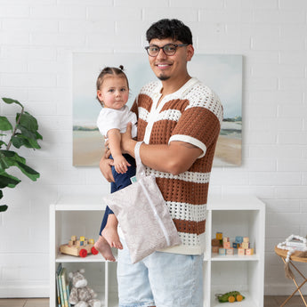 A man in a knit shirt holds a toddler and a Bumkins Wet/Dry Clutch: Wander amid toy shelves and a plant in the backdrop.
