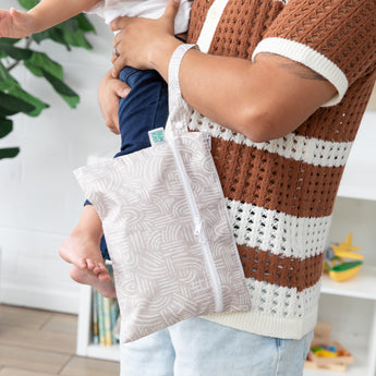 A person holds a child and a Bumkins Wet/Dry Clutch: Wander, dressed in a stylish brown and white knit top and jeans.