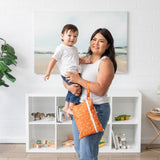 A woman holds a toddler and smiles beside a white brick wall, bookshelf, and plant, holding a Bumkins Wet/Dry Clutch: Grounded on her arm.