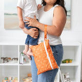 A person in white tank top holds a child and a Bumkins Wet/Dry Clutch: Grounded in a bright room with a bookshelf filled with toys behind.