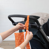 Hands attach the Bumkins Wet/Dry Clutch: Grounded, an orange patterned waterproof bag, to a stroller against a white brick wall.