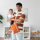 In a living room, a man holds a child and a Bumkins Wet/Dry Clutch: Grounded; white shelves and wall art are in the background.