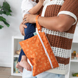 In a cozy room with a bookshelf and plant, a person cradles a baby and holds a Bumkins Wet/Dry Clutch: Grounded on their arm.
