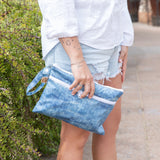 A person in shorts holds a Roamio by Bumkins Wet/Dry Clutch: Crushed Denim by greenery outdoors.