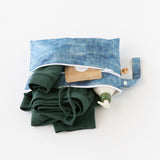 Roamio by Bumkins Wet/Dry Clutch: Crushed Denim lays open on a white background, holding clothes, a lotion bottle, and brown packet.