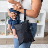A person holding a child and a Bumkins Wet/Dry Clutch: Black, standing in a room with shelves in the background.