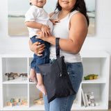 A woman in a toy-filled living room holds a toddler and carries a Bumkins Wet/Dry Clutch: Black, featuring waterproof fabric.