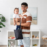 A man with a toddler smiles at the camera in a toy-filled room, holding Bumkins Wet/Dry Clutch: Black on his arm.