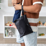 A person carrying a child and a Bumkins Wet/Dry Clutch: Black with waterproof fabric, surrounded by toys and decorations.