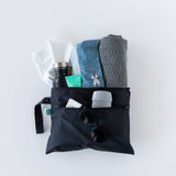 Bumkins Wet/Dry Clutch: Black holds a water bottle, blue clothes, gray towel, toiletries, sunglasses, and deodorant on a white background.